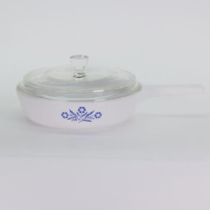 May include: White saucepan with a clear glass lid and handle. The pan features a blue floral design on the side. The lid has a clear knob. This vintage cookware is ideal for cooking.