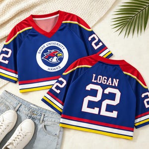 May include: Blue jersey with red and yellow accents. Features the text "BRIAR UNIVERSITY HAWKS" and a hawk logo on the front. The number "22" is on the sleeves and back, with the name "LOGAN" on the back. Paired with jeans and white sneakers.