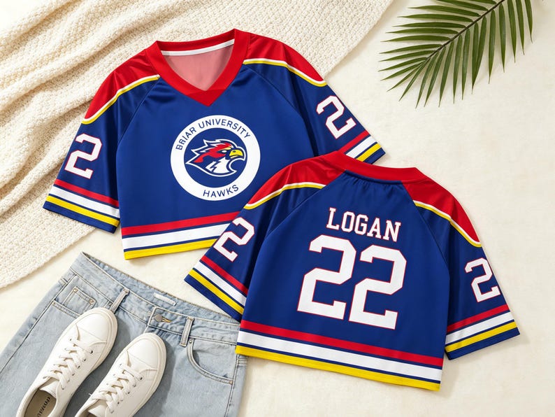 May include: Blue jersey with red and yellow accents. Features the text "BRIAR UNIVERSITY HAWKS" and a hawk logo on the front. The number "22" is on the sleeves and back, with the name "LOGAN" on the back. Paired with jeans and white sneakers.