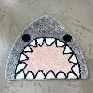 May include: A gray shark-shaped bath mat with a pink interior mouth, white teeth, and black accents. The rug is designed to resemble a shark's open mouth, adding a playful touch to any bathroom decor.