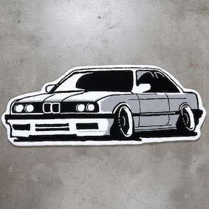 Peut inclure: Un tapis en forme de voiture gris et blanc avec un contour noir. Le tapis présente une illustration détaillée d'une voiture classique, comprenant des phares, une calandre et des roues. La voiture est représentée de profil, en mettant l'accent sur son design.