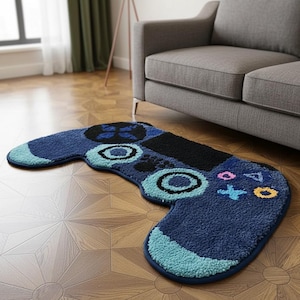 Puede incluir: Una alfombra azul, negra y turquesa con forma de mando de videojuegos. La alfombra presenta diseños de botones circulares y en forma de cruz, con un borde turquesa. Está colocada sobre un suelo de madera cerca de un sofá gris.