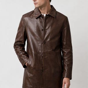 May include: A dark brown leather coat featuring a classic collar, a button-front closure with five visible buttons, long sleeves, and two side pockets. The coat has a smooth, polished finish and a structured fit that extends to the mid-thigh area.
