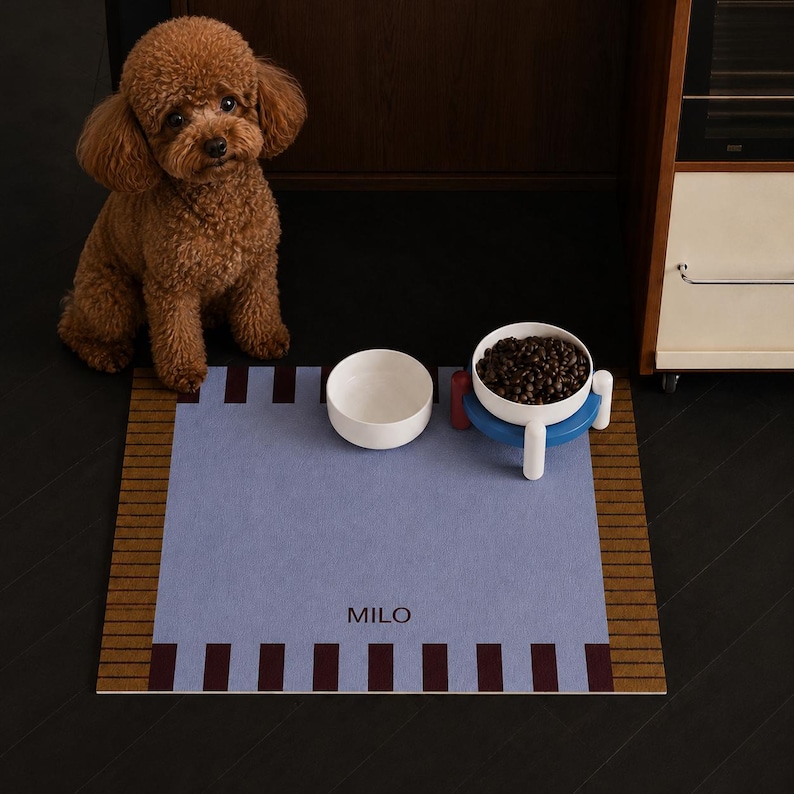 May include: A rectangular pet mat in light blue with a brown striped border and dark brown rectangular accents. The name MILO is printed in dark brown letters on the mat. Two white pet bowls are placed on the mat, one filled with brown kibble.