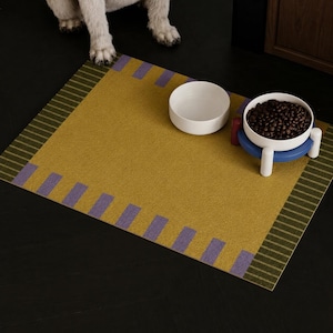 May include: A rectangular mustard yellow pet mat featuring a border with purple rectangular accents and dark green striped edges. Two white bowls are placed on the mat, one empty and one filled with dark brown dry pet food on a blue stand.