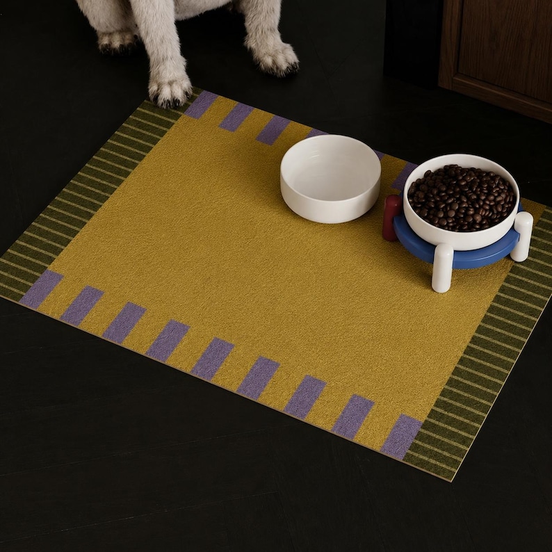 May include: A rectangular mustard yellow pet mat featuring a border with purple rectangular accents and dark green striped edges. Two white bowls are placed on the mat, one empty and one filled with dark brown dry pet food on a blue stand.
