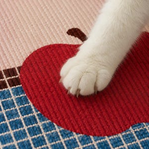 May include: A textured fabric mat featuring a large red apple graphic with a brown stem, set against a background of light pink and a blue grid pattern. A white cat paw with visible claws rests on the surface of the red apple design.