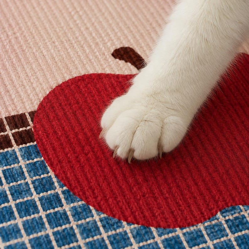 May include: A textured fabric mat featuring a large red apple graphic with a brown stem, set against a background of light pink and a blue grid pattern. A white cat paw with visible claws rests on the surface of the red apple design.