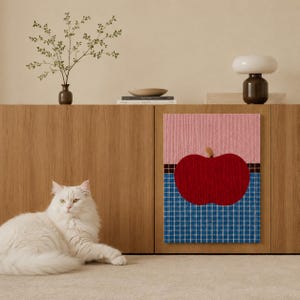 May include: A rectangular wall art piece featuring a large red apple graphic centered on a background divided into a pink textured upper section and a blue and white grid patterned lower section, separated by a thin black and brown checkered border.