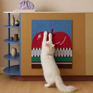 May include: A white cat stands on its hind legs, reaching up to touch a rectangular wall hanging. The hanging features a large red apple graphic against a blue background, with a colorful geometric pattern at the bottom and a white star accent.