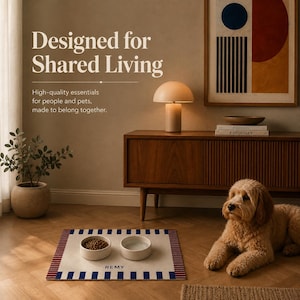 May include: A rectangular pet mat with a cream center, blue and red striped borders, and the name REMY printed in black. Two round ceramic bowls, one filled with dry food and one with water, sit on the mat. A golden-colored dog rests nearby.