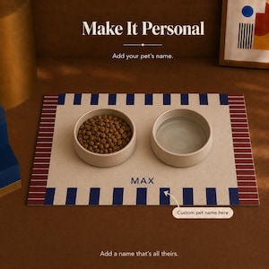 May include: A rectangular pet feeding mat in beige with dark blue vertical stripes along the top and bottom edges and maroon striped borders on the sides. Two round, light-colored bowls sit on the mat, one filled with brown kibble and one with water. The name MAX is printed in blue text on the mat.