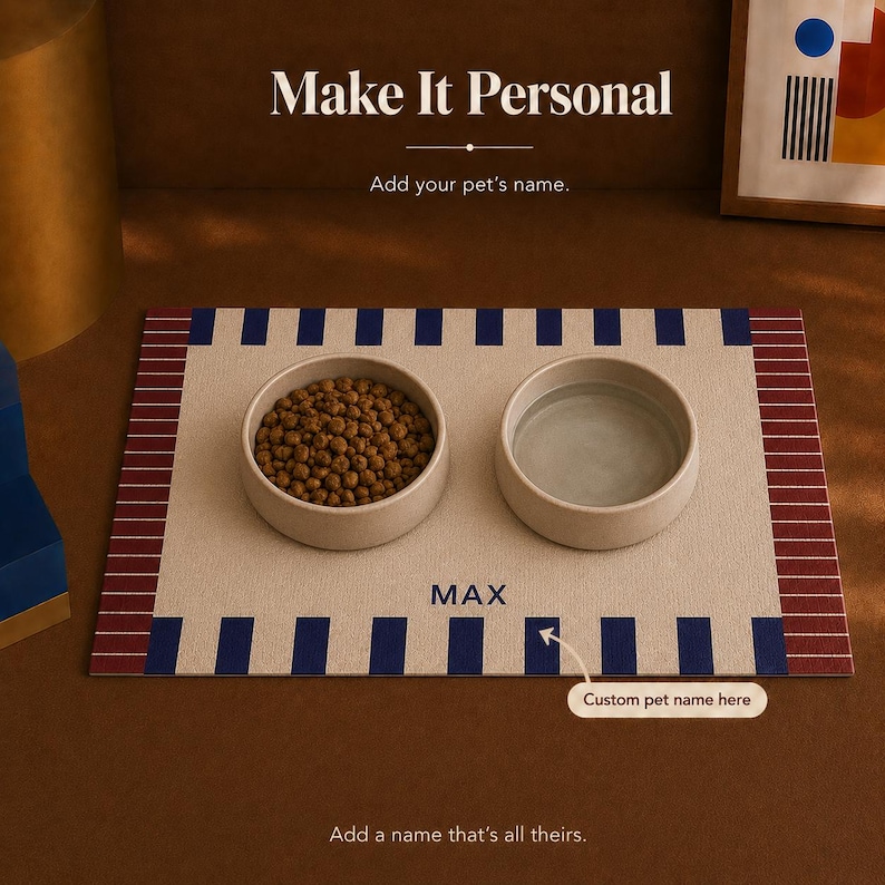May include: A rectangular pet feeding mat in beige with dark blue vertical stripes along the top and bottom edges and maroon striped borders on the sides. Two round, light-colored bowls sit on the mat, one filled with brown kibble and one with water. The name MAX is printed in blue text on the mat.