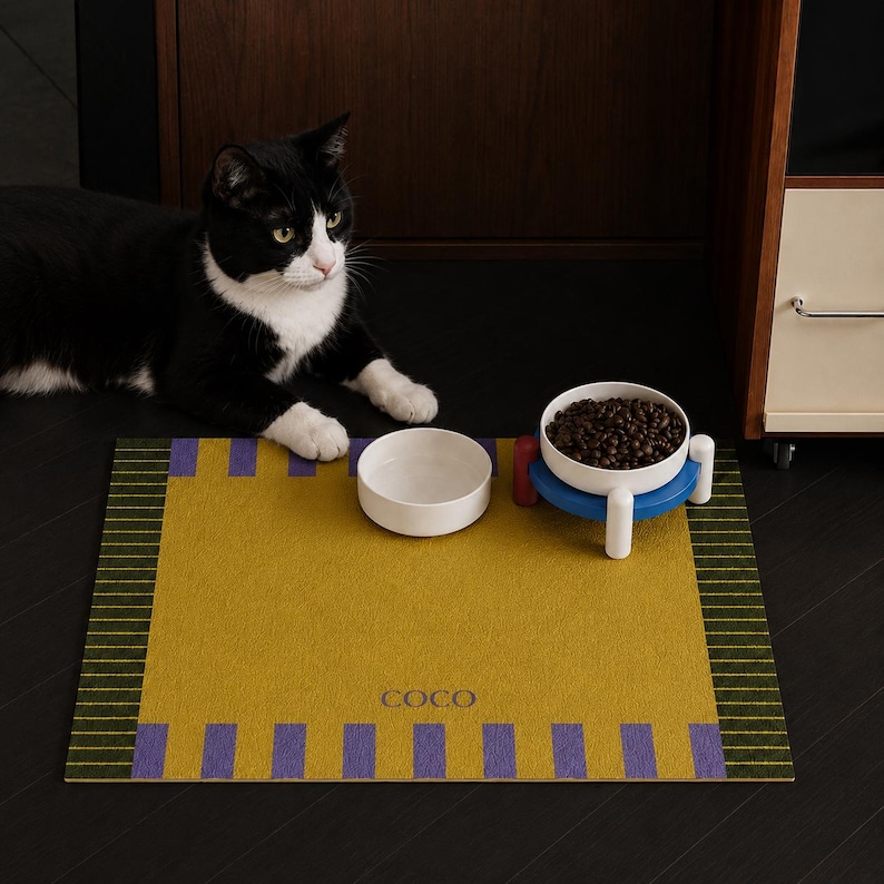May include: A rectangular mustard yellow pet mat with purple vertical stripes on the top and bottom edges and dark green striped borders on the sides. The mat features the name COCO printed in the center, with two white bowls placed on top.