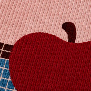 May include: A corduroy fabric featuring a large, deep red apple graphic with a dark brown stem. The background is a soft, light pink corduroy, and a portion of a blue and white grid pattern is visible along the bottom left edge of the textile.
