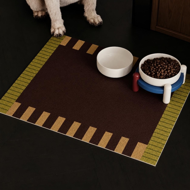 May include: A rectangular pet feeding mat with a dark brown center, bordered by olive green stripes and tan rectangular accents. Two white ceramic bowls sit on the mat, one empty and one filled with brown kibble, supported by a blue and red stand.