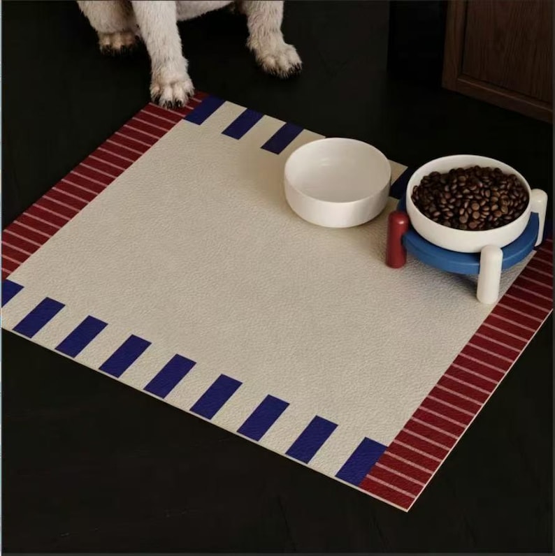 May include: A rectangular pet feeding mat with a textured cream center, featuring a border of red stripes on two sides and a pattern of blue rectangular blocks along the other edges. Two white ceramic bowls sit on the mat, one filled with brown kibble.