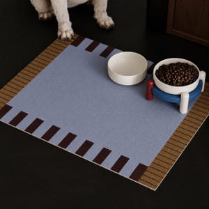 May include: A square pet feeding mat with a textured light blue center, brown striped borders, and dark brown rectangular accents. Two white ceramic bowls sit on the mat, one empty and one filled with brown dry kibble on a blue stand.