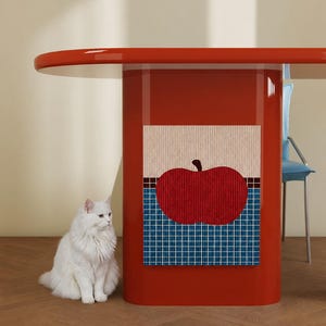 May include: A glossy red table with a central pedestal base featuring a square wall art piece depicting a large red apple against a blue grid background. A fluffy white cat sits on the floor next to the table, which has a smooth, rounded edge.