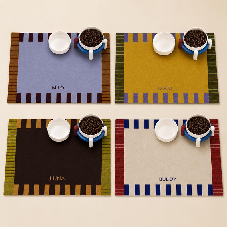 May include: Four rectangular pet feeding mats with striped borders, each featuring a personalized name: MILO, COCO, LUNA, and BUDDY. Each mat includes a white ceramic bowl and a raised blue bowl stand filled with dark brown kibble.