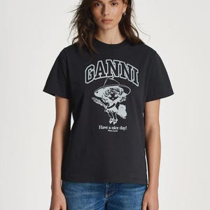 May include: Black crew neck t-shirt with the word "GANNI" in white block letters above a cartoon graphic. The graphic features a girl wearing a hat and holding flowers. The text "Have a nice day!" is below the graphic.