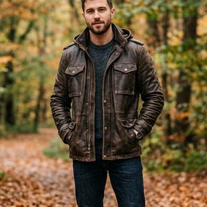 May include: A brown leather jacket with a hood, worn over a gray sweater and dark blue jeans. The jacket has multiple pockets and a button-up front. The person is standing outdoors in a forest setting with autumn leaves.