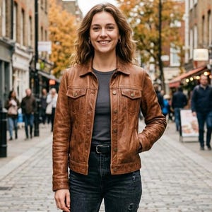 May include: A brown leather jacket with a classic design, featuring two chest pockets and a button-up front. The jacket is worn with a gray shirt and black jeans. The model is smiling in an outdoor setting.