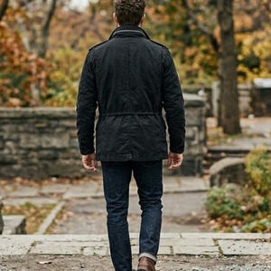 K&ouml;nnte beinhalten: Eine Person in einer schwarzen Jacke, dunkelblauen Jeans und braunen Stiefeln geht auf einem Steinweg weg. Die Jacke hat eine Taille mit Kordelzug und einen Kragen. Der Hintergrund zeigt B&auml;ume und eine Steinmauer.