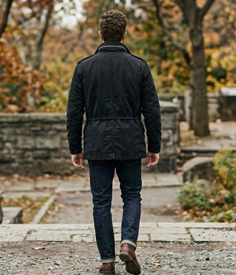 K&ouml;nnte beinhalten: Eine Person in einer schwarzen Jacke, dunkelblauen Jeans und braunen Stiefeln geht auf einem Steinweg weg. Die Jacke hat eine Taille mit Kordelzug und einen Kragen. Der Hintergrund zeigt B&auml;ume und eine Steinmauer.
