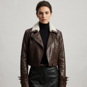 May include: A dark brown leather jacket featuring a white faux fur collar, a diagonal front zipper closure, and decorative petal-like leather accents at the cuffs. The jacket is worn over a black ribbed turtleneck sweater and a black skirt.