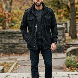 May include: A black jacket with multiple pockets, a grey jumper, and dark blue jeans. The jacket has a zip closure and a collared neckline. The person is wearing brown leather boots. The background features a stone wall and trees.
