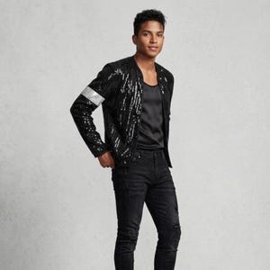 Può includere: Un giubbotto bomber nero con paillettes caratterizzato da una fascia argentata sulla manica destra, indossato sopra una canotta nera a girocollo e jeans neri strappati. La giacca presenta un design senza colletto e una chiusura a zip.