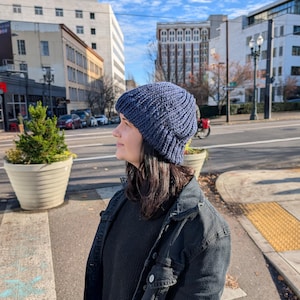 May include: A navy blue knit beanie with a ribbed design. The hat is worn by a person wearing a black sweater and a black denim jacket. The background shows a city street with buildings and trees.