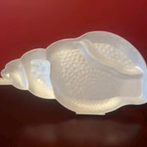 May include: A white, seashell-shaped serving dish with a textured surface. The dish has a wide, open design and a smooth, curved edge. A small tag is attached to the dish. The background is a dark red.