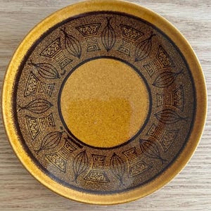 May include: A round ceramic plate in brown and yellow. The plate has a central yellow circle and a brown border with a repeating geometric and leaf-like pattern. The outer rim is a darker brown, creating a contrasting edge.