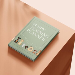 Peut inclure: Un agenda d'entraînement pour chiots vert sauge avec le texte "PUPPY TRAINING PLANNER" sur la couverture. La couverture présente des illustrations de diverses races de chiens. L'agenda est sur une surface couleur pêche.