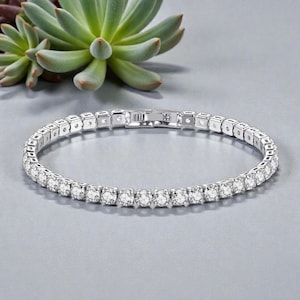 Puede incluir: Una pulsera de tenis plateada con piedras preciosas redondas y transparentes engastadas en un marco cuadrado. La pulsera se muestra sobre una superficie gris claro, con una planta suculenta en el fondo.