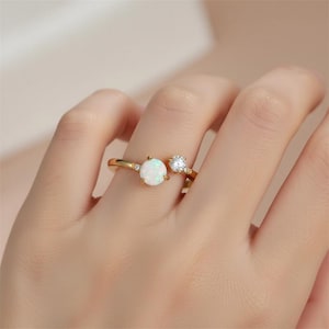 May include: A gold ring featuring a large, round, white opal and a smaller, clear gemstone. The ring has a delicate design with small accent stones on the band. The ring is shown on a person's finger.
