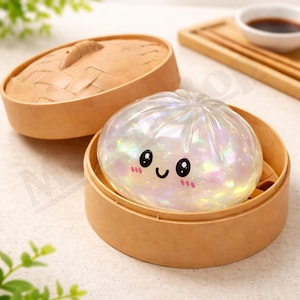 Puede incluir: Un objeto perlado y sonriente con forma de bollo bao se encuentra dentro de un vaporizador de bambú. El bollo tiene una cara linda con ojos negros y una boca curvada, y mejillas rosadas. El vaporizador tiene tapa y es de bambú marrón claro.