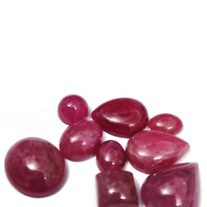 May include: A collection of polished, cabochon-cut gemstones in varying shapes and sizes. The gems are a deep, rich red color, with a smooth, reflective surface. The arrangement includes oval, teardrop, and rectangular stones.