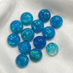 Può includere: Un gruppo di opali rotondi e lucidi, in varie tonalità di blu e verde acqua. Gli opali hanno una finitura lucida e mostrano lampi di colore interni. Sono disposti su un tessuto bianco e strutturato.