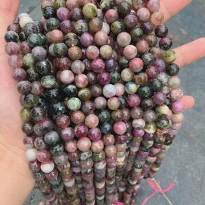 May include: Strands of multi-colored tourmaline gemstone beads. The beads are round, with a mix of pink, green, brown, and grey hues. The beads are strung together, ready for jewelry making.