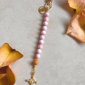 May include: A rosary keychain with pink and white beads, a gold-coloured cross pendant, and a gold-coloured clasp. The keychain also features a small gold charm. The text "REVERIE ROSARIES" is visible in the background.