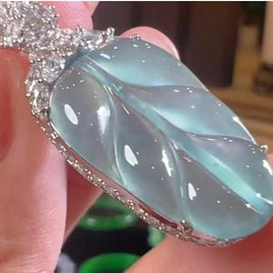 Puede incluir: Un colgante en forma de hoja con una piedra preciosa translúcida de color azul claro. El colgante está engastado en un marco de metal plateado, con un grupo de pequeñas piedras transparentes en la parte superior. La piedra preciosa tiene vetas visibles y el diseño general es elegante.