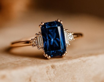May include: A gold ring featuring a large, rectangular, deep blue gemstone as the centerpiece. Two smaller, rectangular, clear gemstones flank the main stone. The ring is set on a neutral-toned surface, with a blurred background.