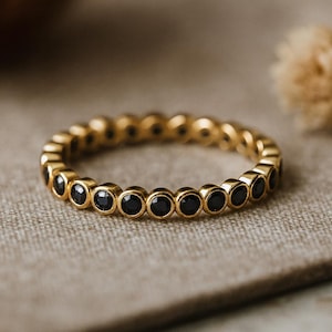 May include: A gold-colored ring with a band of small, round, black gemstones. The ring is set on a neutral-toned fabric, showcasing the jewelry's design and color. The ring is a piece of fashion jewelry.
