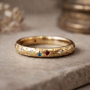 Op de afbeelding: Een gouden ring met een sterren- en stippenpatroon, met een blauwe, rode en twee witte edelstenen. De ring ligt op een neutrale ondergrond en toont het ingewikkelde ontwerp en vakmanschap. De ring is een sieraad.