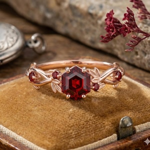 May include: A rose gold ring with a hexagonal, deep red gemstone centre. The ring features delicate leaf and berry accents on the band, with smaller red stones. The ring is displayed on a velvet surface.