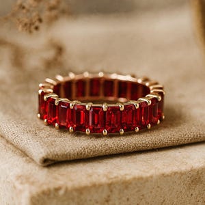 May include: A gold-toned ring featuring a continuous band of rectangular, vibrant red gemstones. The ring is set on a neutral-toned fabric, showcasing the rich color and design. The gemstones are evenly spaced, creating a bold and elegant look.