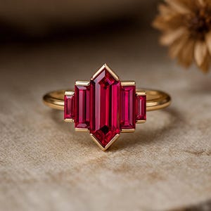 Peut inclure: Bague en or avec un grand rubis central taillé en rectangle, flanqué de deux petits rubis rectangulaires de chaque côté. Le design de la bague est géométrique et d'inspiration Art Déco, avec un anneau en or poli.